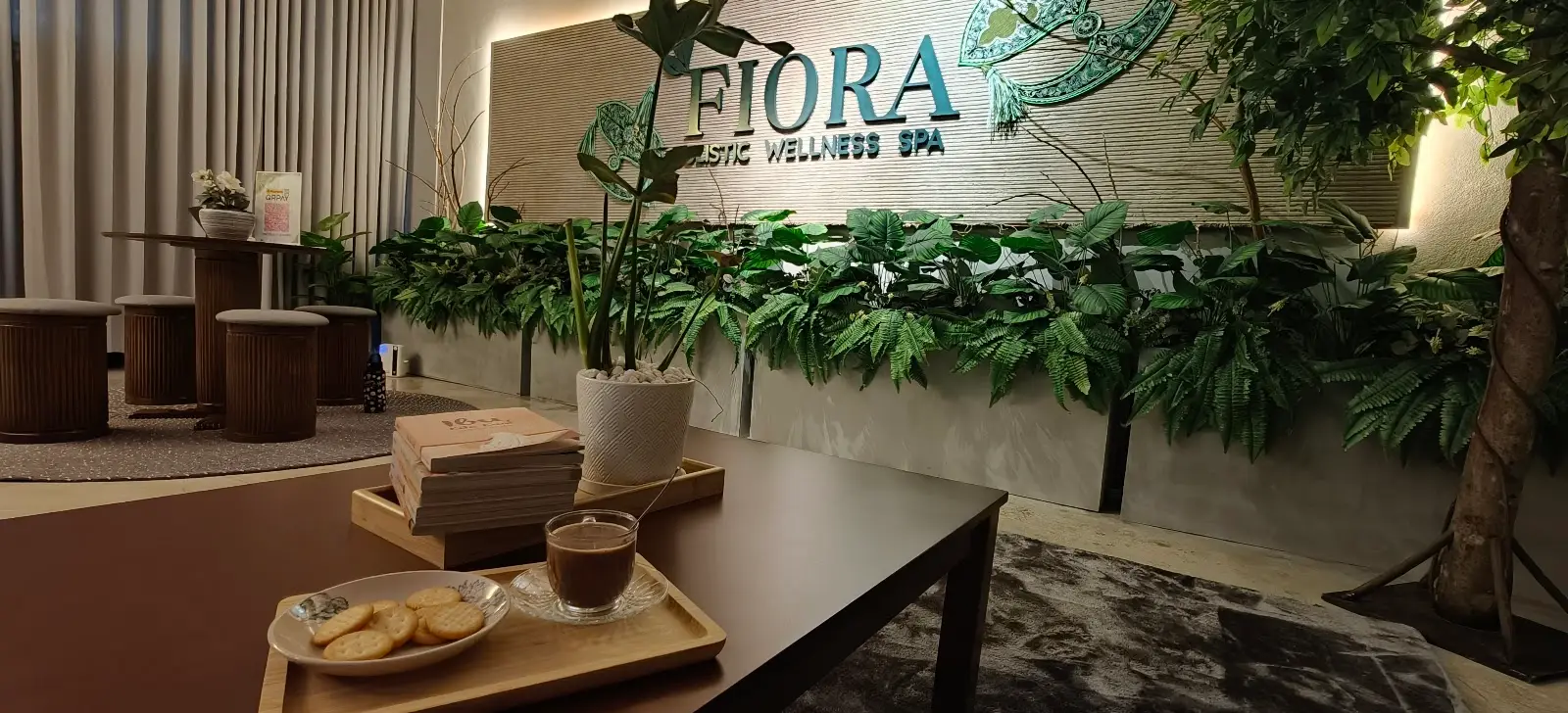 Ruang menunggu FIORA dengan dinding logo, tumbuhan hijau dan dulang air serta biskut untuk pelanggan